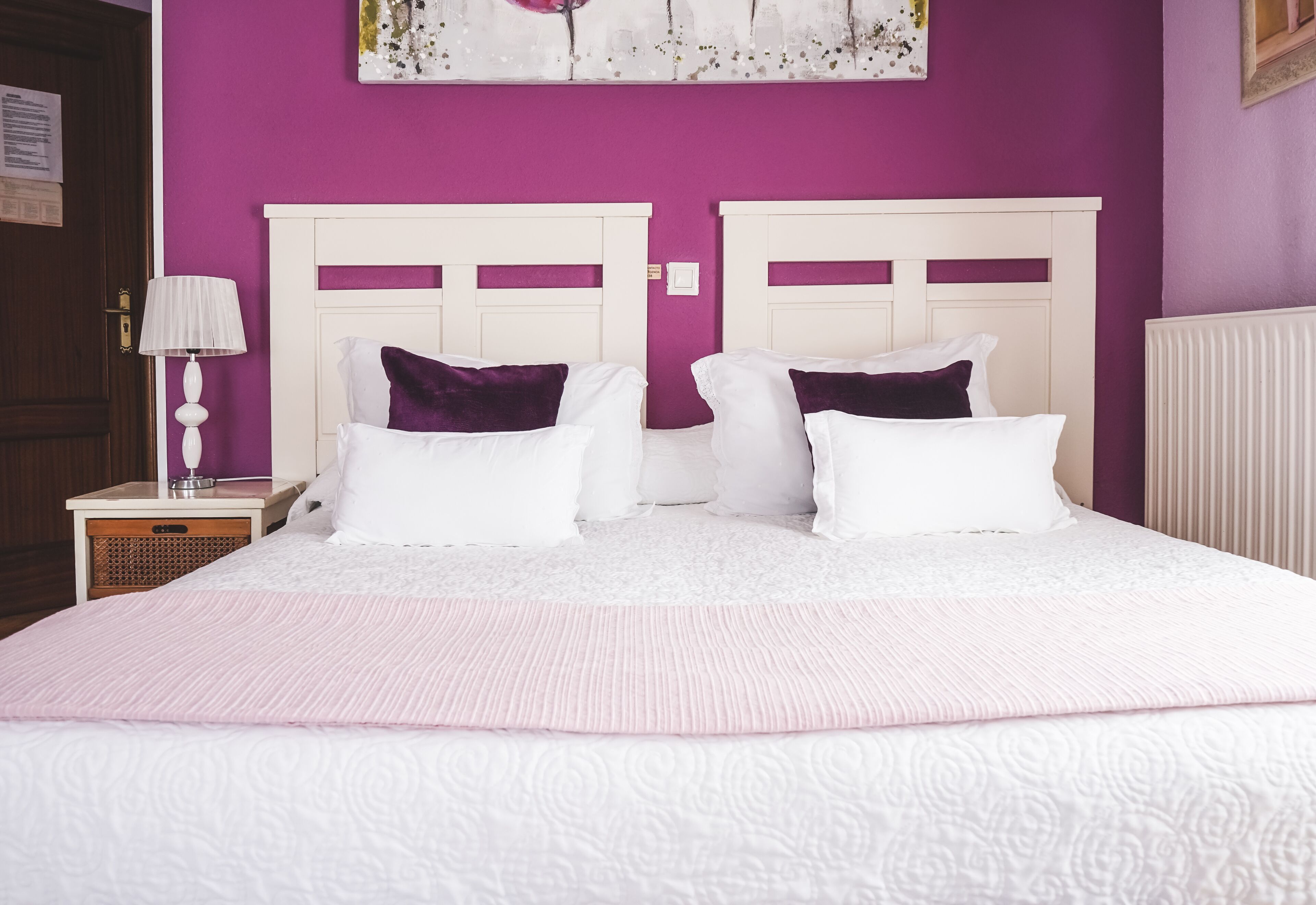 Una cama doble con almohadas blancas y moradas, cabecera blanca y colcha rosa en una habitación con paredes moradas.