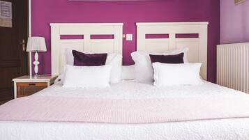 Una cama doble con almohadas blancas y moradas, cabecera blanca y colcha rosa en una habitación con paredes moradas.