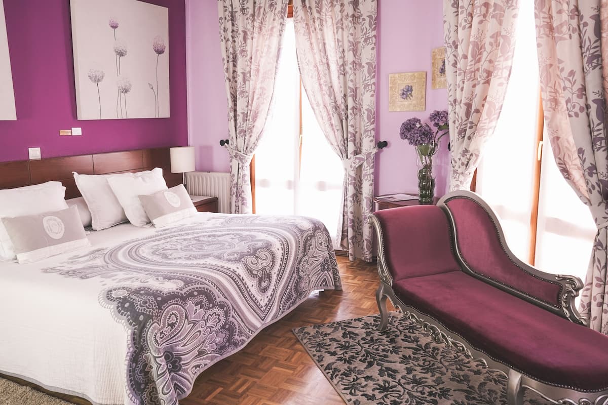 A bedroom with a large bed, a red armchair, a wooden floor, and a window with curtains.