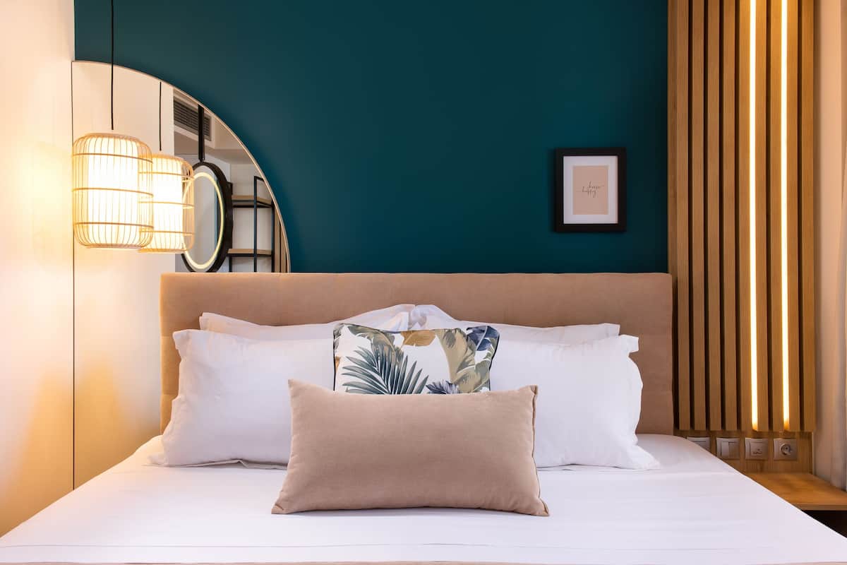 A neatly made bed with white and beige pillows, a large mirror, and a wall-mounted light fixture.