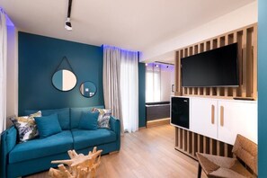 A modern living room with a teal sofa, a wooden coffee table, a flat-screen TV mounted on a wooden panel, and a dining area with a white cabinet and a small table. - SKS Luxury Suites & Rooms (Katerini)