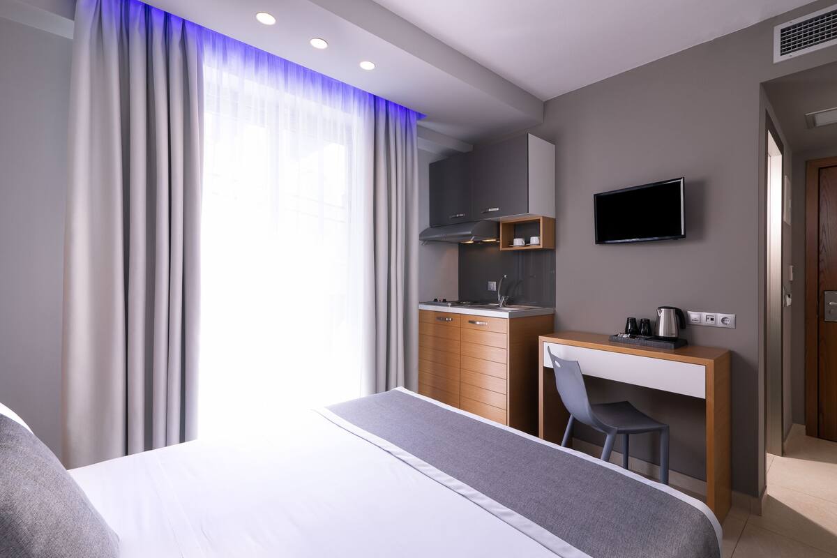 A modern hotel room with a bed, a desk, a chair, a television, and a kitchenette.