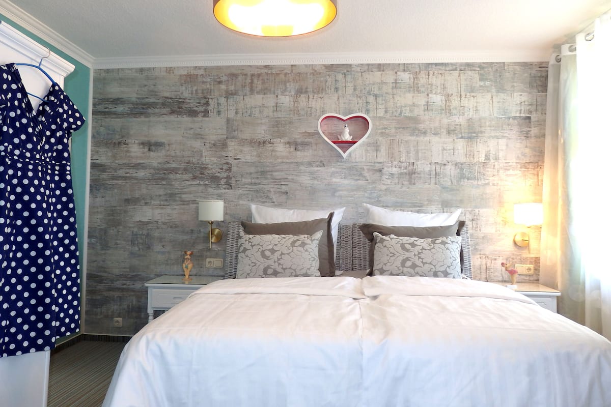 A bedroom with a bed, a nightstand, a wall-mounted heart-shaped decoration, and a polka dot dress hanging on a door.