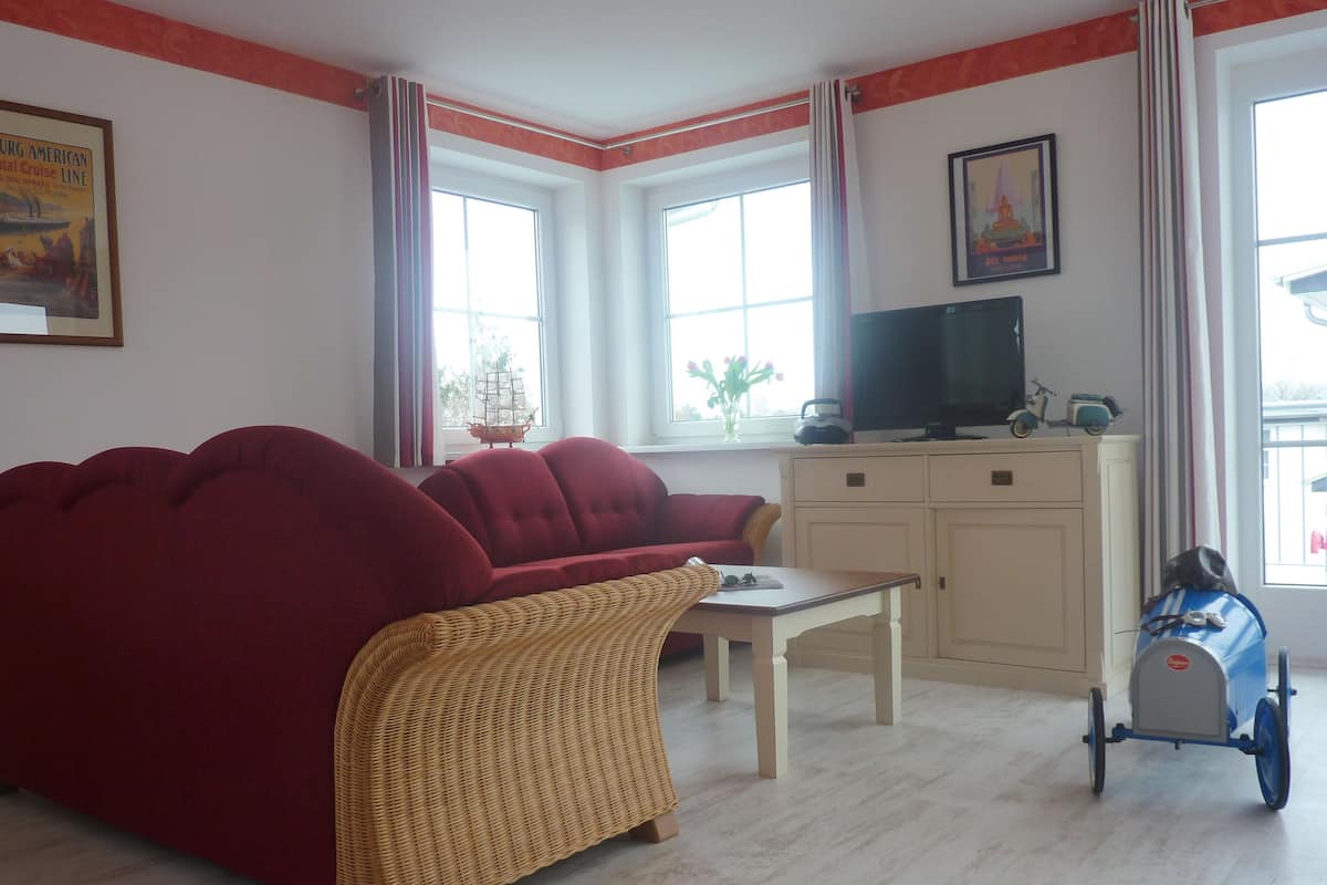 A living room with a red sofa, a wicker chair, a small table, a television on a cabinet, and a cart with a gas cylinder.
