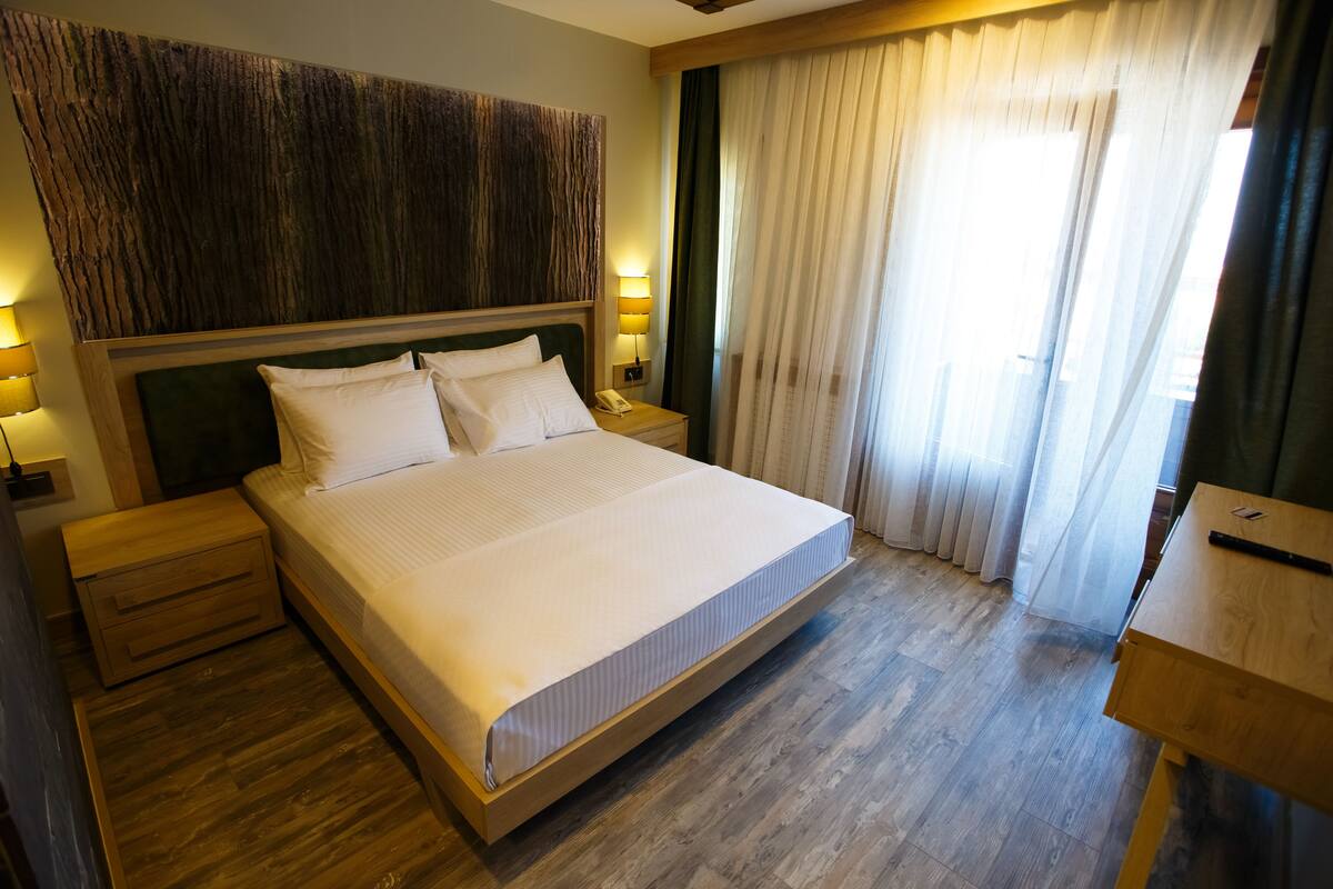 A hotel room with a large bed, wooden headboard, bedside tables, a nightstand, and a wooden desk.