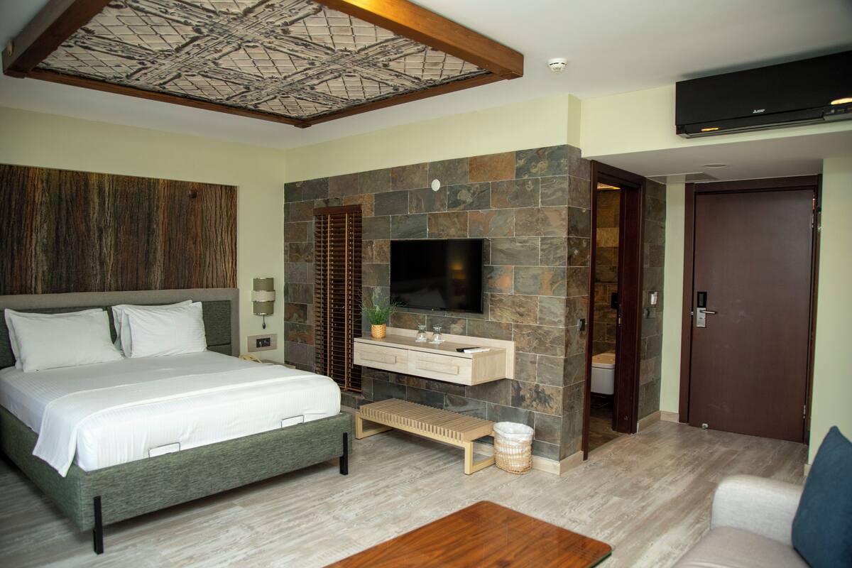 A bedroom with a bed, a TV, a stone wall, a wooden bench, and a door.