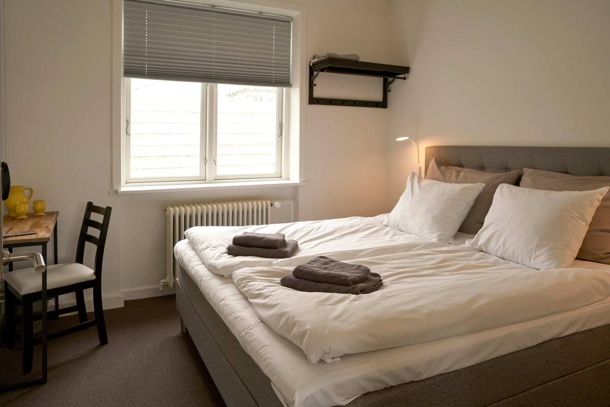 A bedroom with a bed, a chair, a table, a window with blinds, and a radiator.