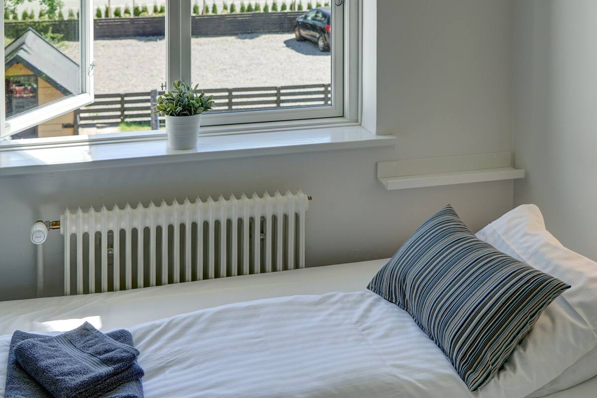 A neatly made bed with white linens and a striped pillow, a radiator below the window, and a small potted plant on the windowsill.