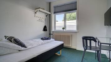 A single bed with a black frame, a small table with a chair, a radiator, a window with blinds, and a wall-mounted TV.