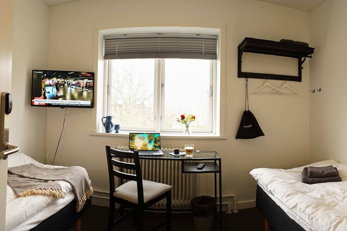A hotel room with a bed, a desk with a laptop, a chair, a radiator, a TV mounted on the wall, and a window with a view of trees.