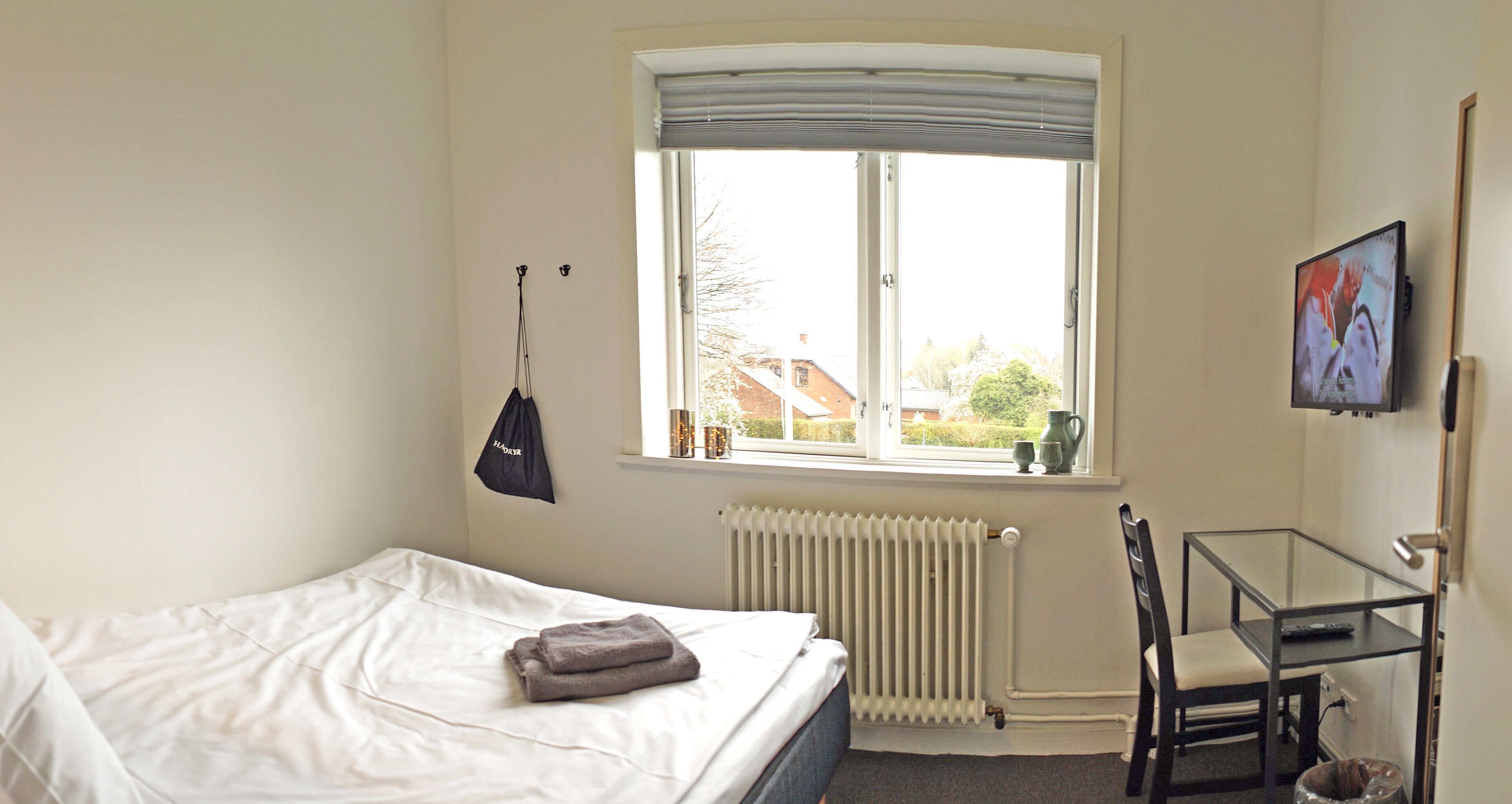 Un dormitorio con cama, ventana con vistas, radiador, una silla y un cuadro en la pared.