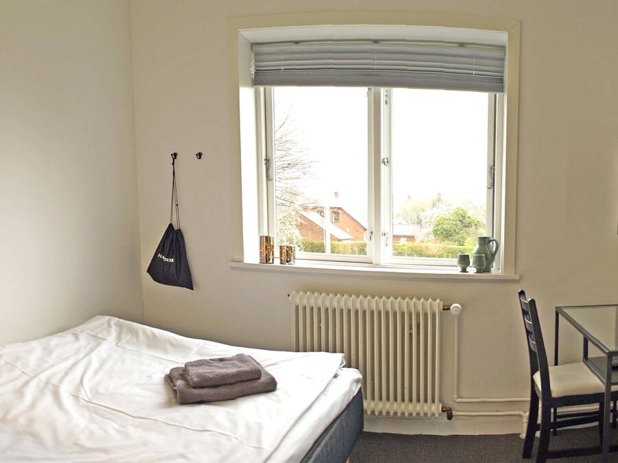 Un dormitorio con cama, ventana con vistas, radiador, una silla y un cuadro en la pared.