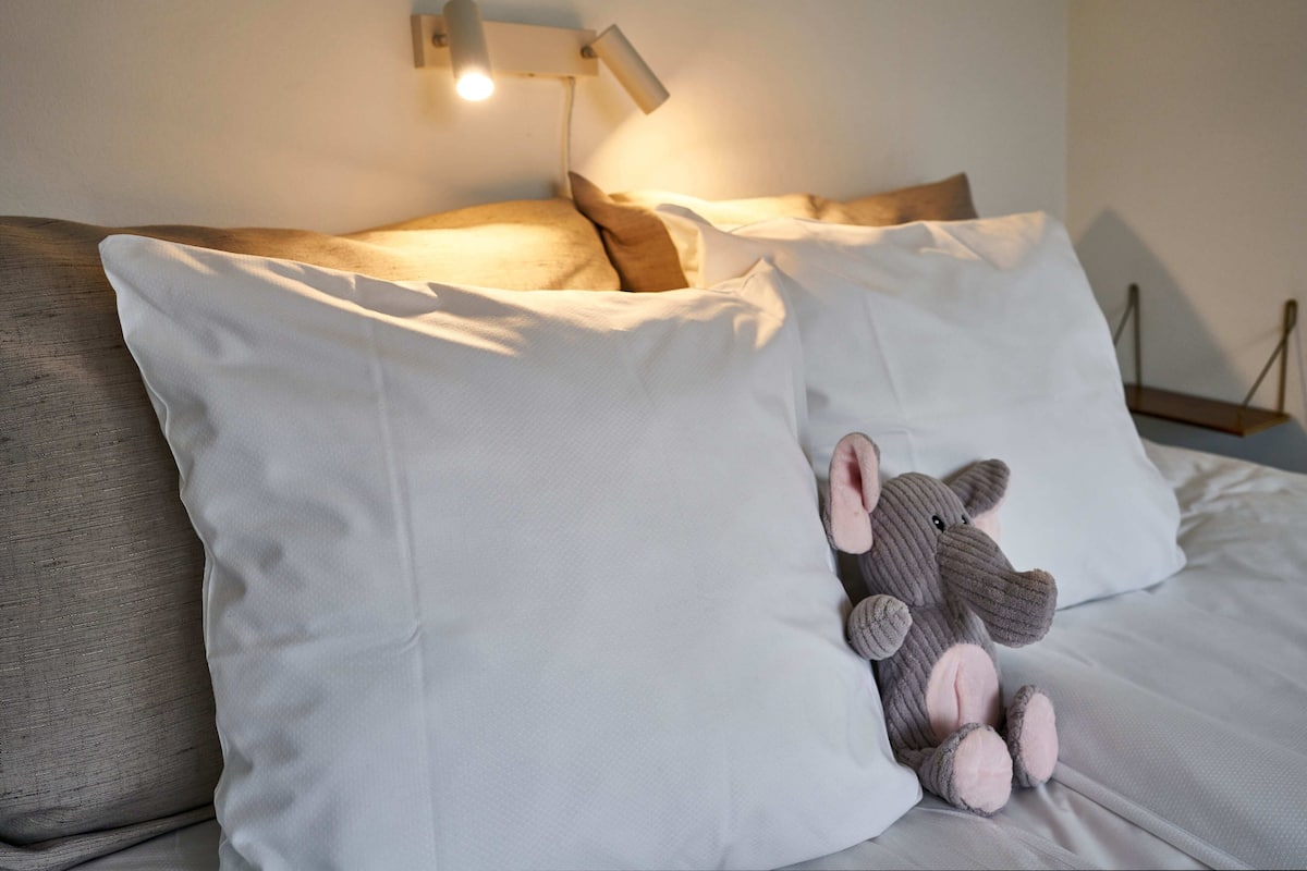 A bed with white and grey bedding, a small stuffed animal, and a wall-mounted light fixture.