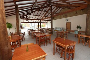 A spacious indoor dining area with wooden tables and chairs, a high ceiling with wooden beams, and large windows allowing natural light. - Recanto das Carpas (Ilhabela)