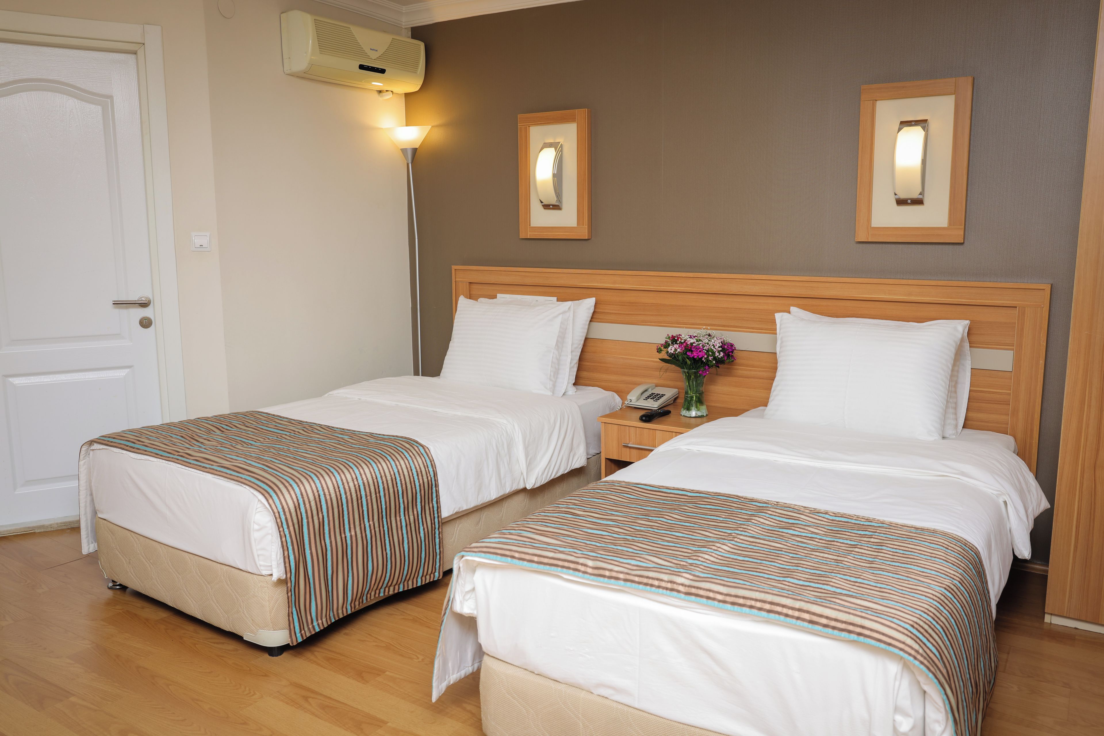 Habitación de hotel con dos camas, una mesita de noche, una lámpara y un aire acondicionado instalado en la pared.
