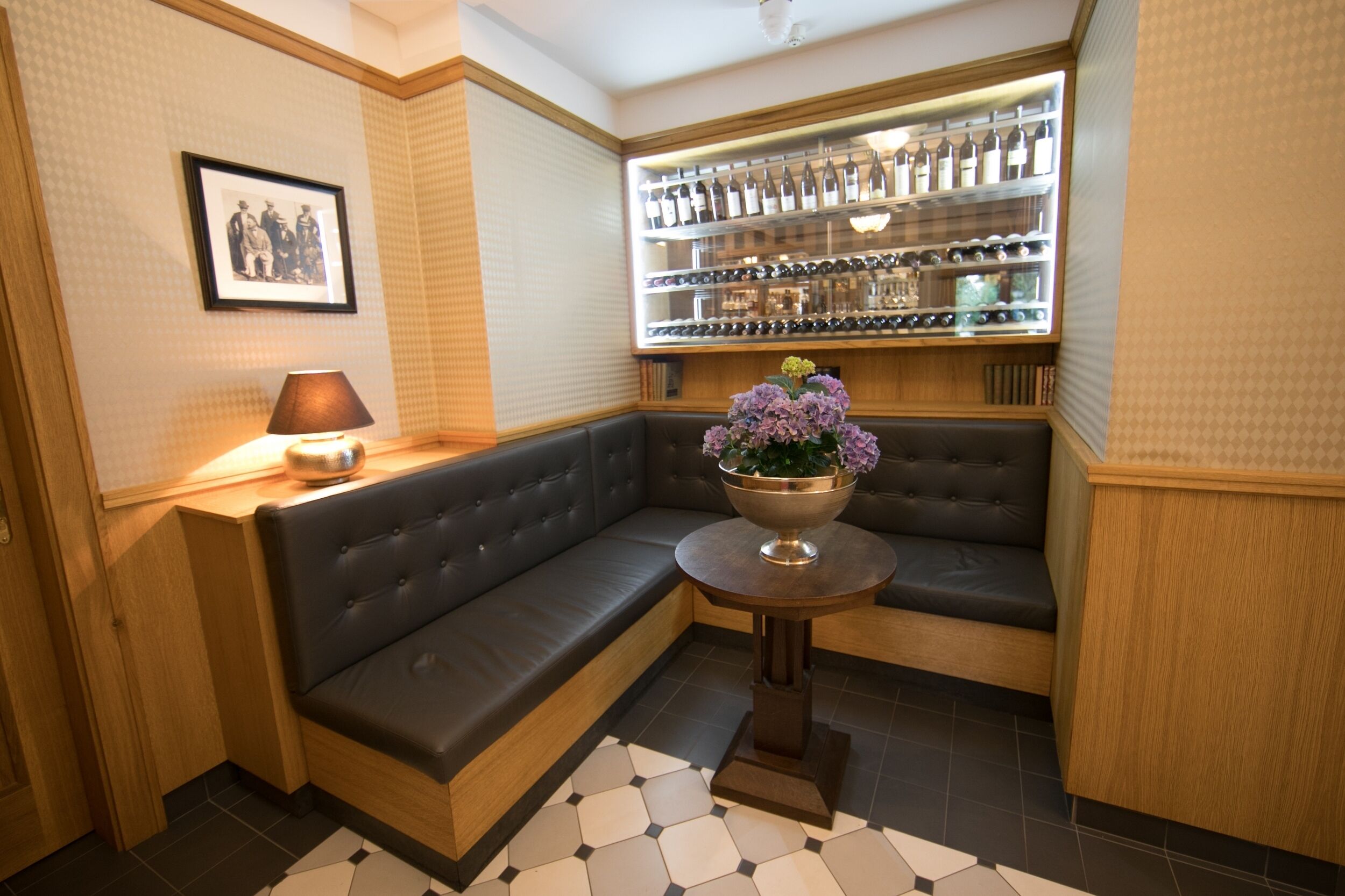 Un coin salon avec un canapé, une petite table ornée d’un vase de fleurs, une étagère murale avec des bouteilles et un tableau encadr é.