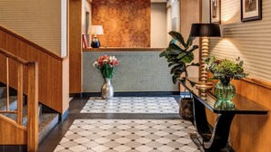 A hotel lobby with a checkered floor, wooden reception desk, and a wall-mounted clock. - Hotel Golfi (Podebrady)