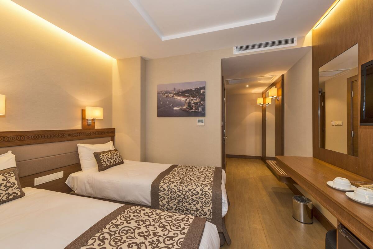 A hotel room with two beds, a wooden headboard, a large mirror, a picture of a cityscape, a wooden bench, and a trash bin.