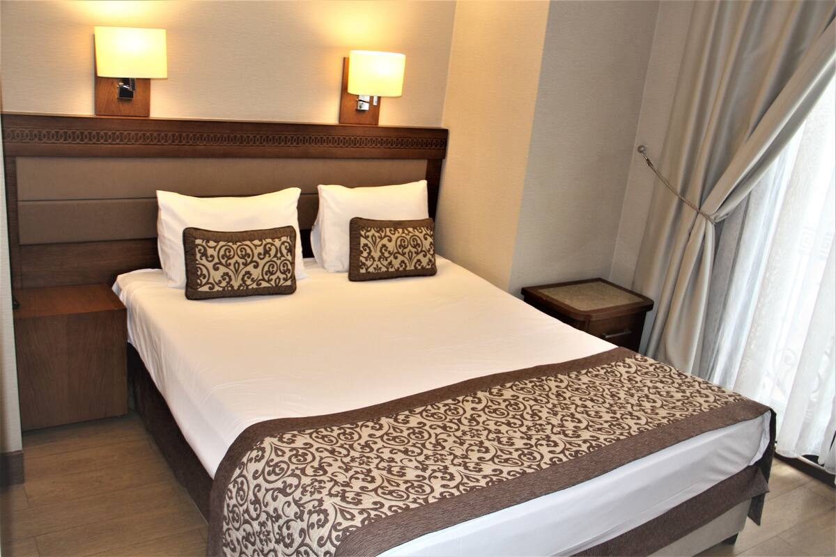 A hotel room with a large bed, two bedside lamps, a nightstand, and a curtain.