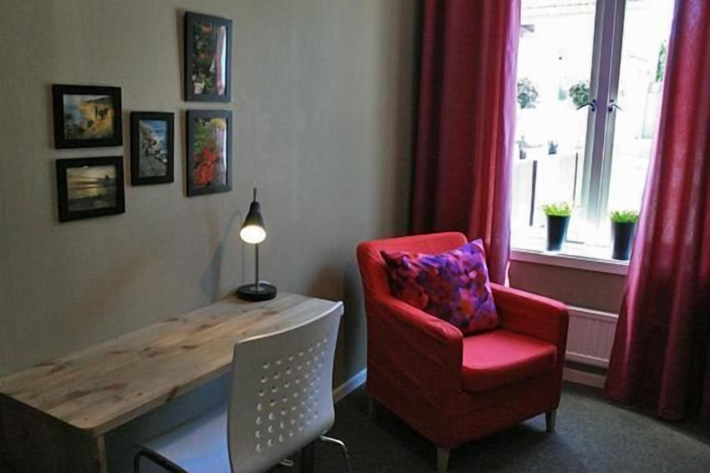 Een kamer met een bureau, een rode stoel met een kleurrijk kussen, een wandlamp en een raam met gordijnen.