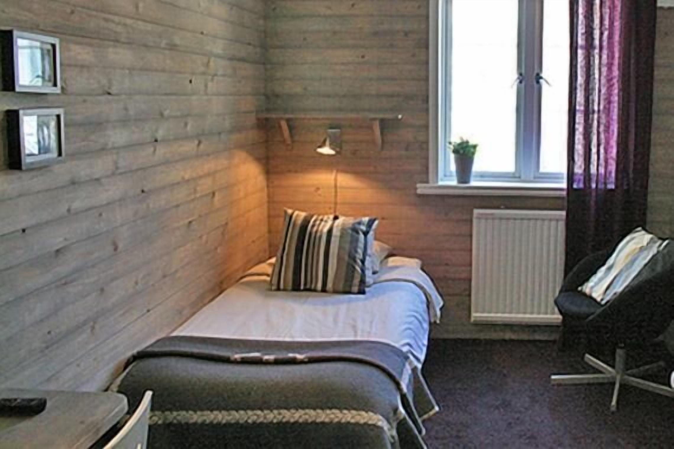 Een kleine, gezellige kamer met houten wanden, een bed met gestreepte kussens, een bedlamp, een stoel en een raam met gordijnen.