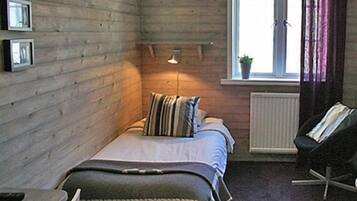 Een kleine, gezellige kamer met houten wanden, een bed met gestreepte kussens, een bedlamp, een stoel en een raam met gordijnen.