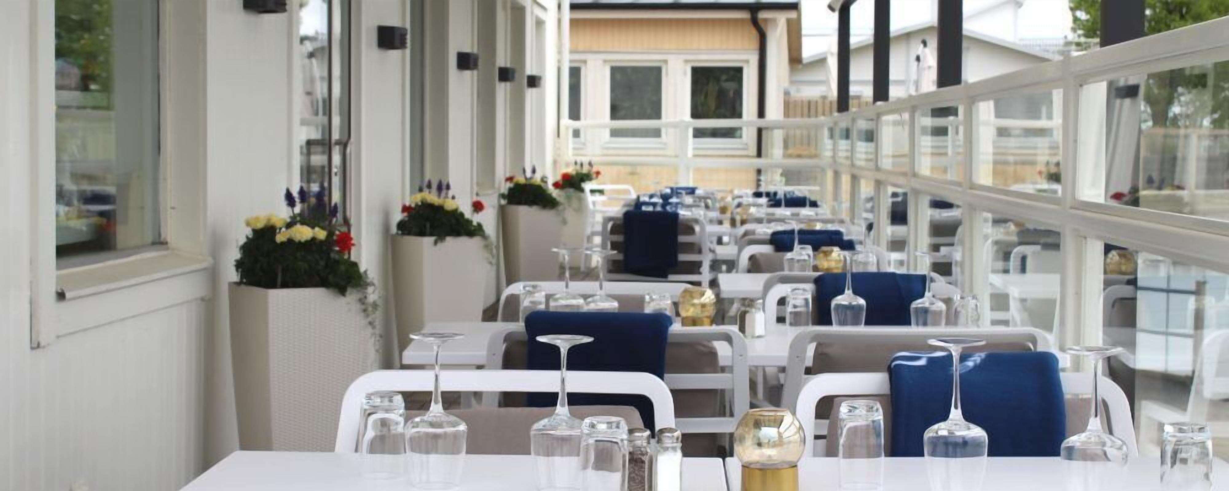 En restaurang med vita bord och blå stolar, placerade i rader med stora glasfönster på ena sidan.