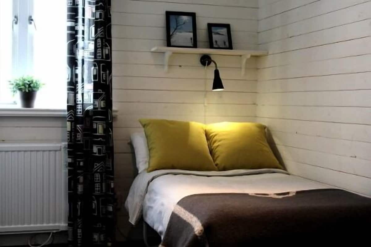 Um quarto pequeno e acolhedor com uma cama, uma janela com cortinas, um radiador e uma estante com duas fotos.