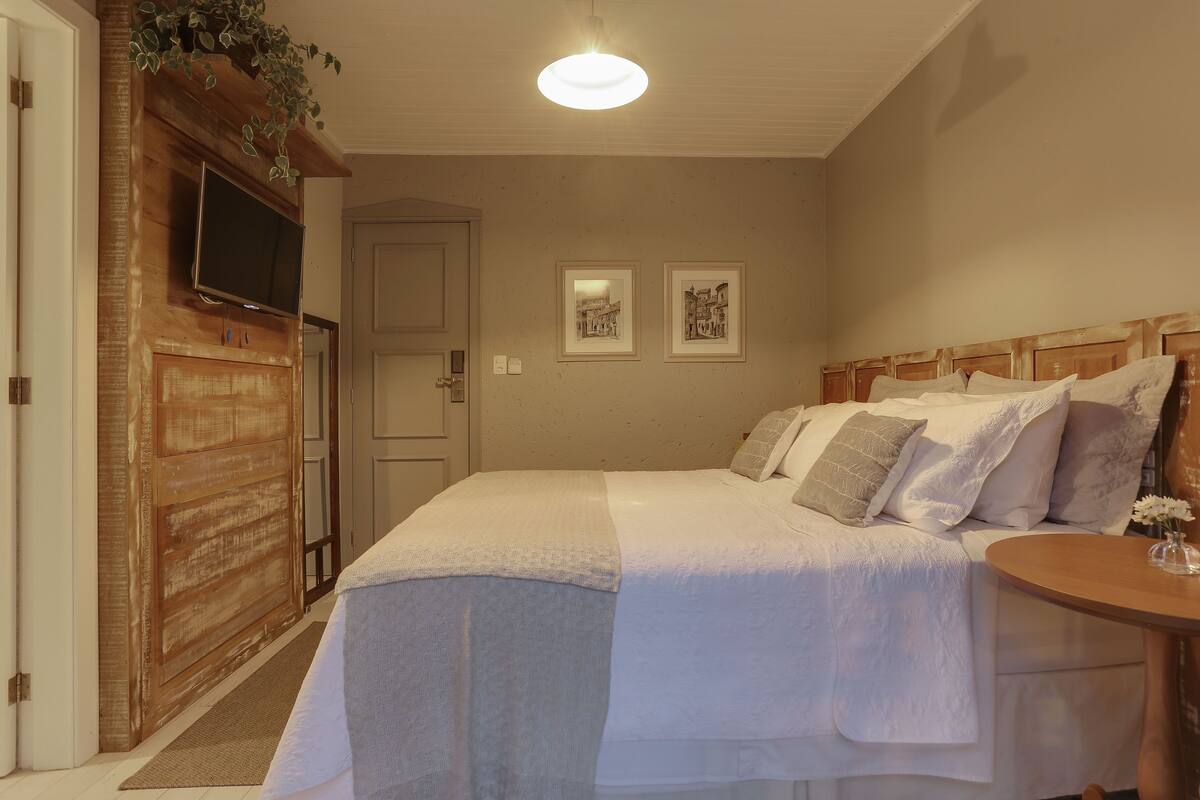 Une chambre à coucher avec un lit, une tête de lit en bois, une table de chevet avec un vase et un téléviseur fixé au mur.