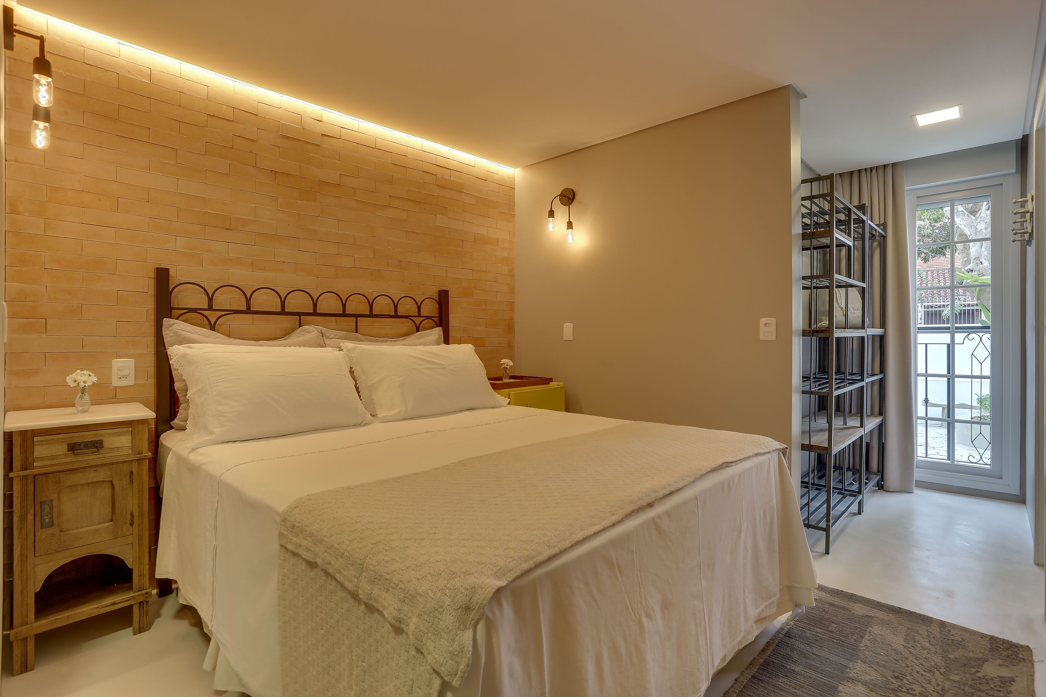A bedroom with a wooden headboard, a bed with white bedding, a nightstand with a vase, and a view of a balcony with a metal railing.