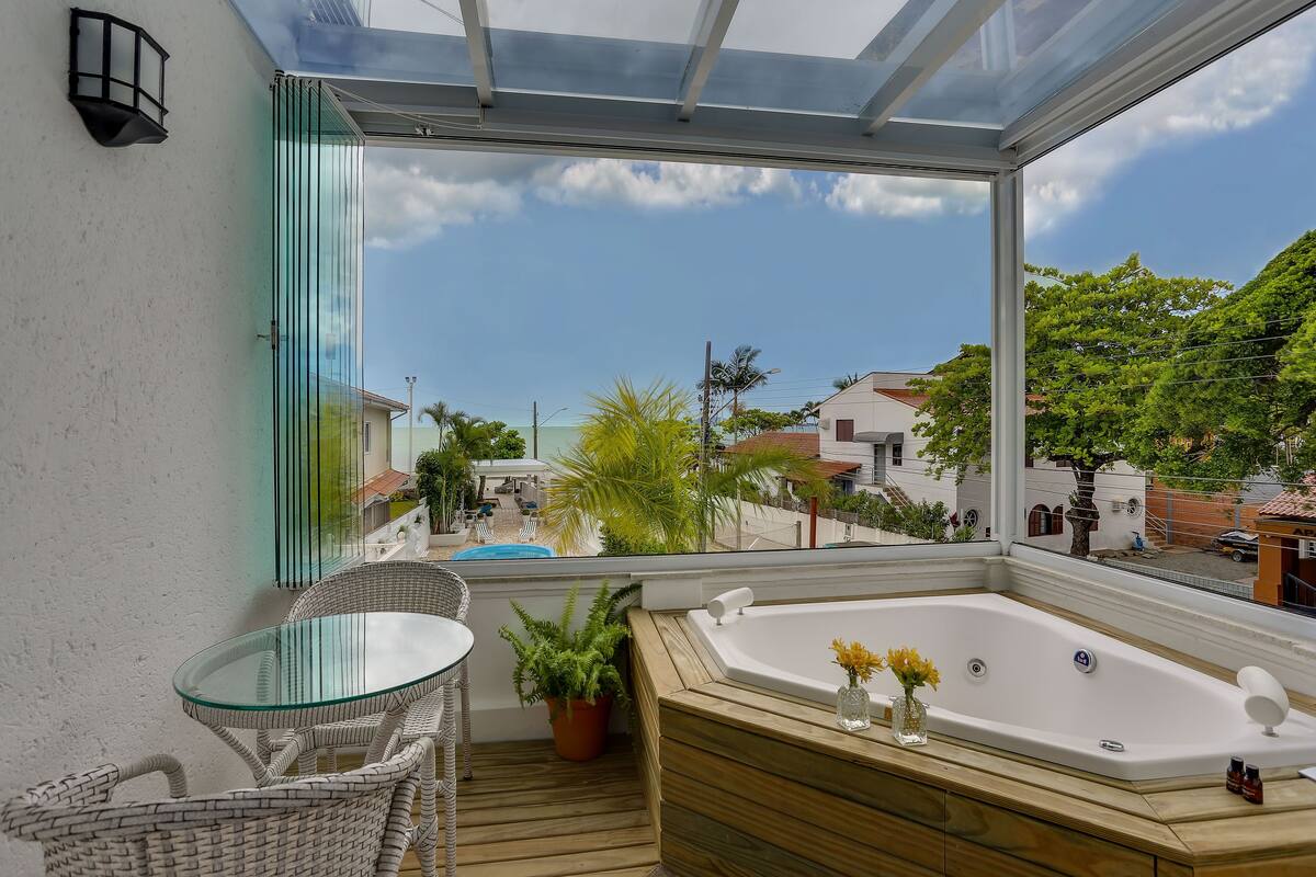 Un balcon avec une porte vitrée, une terrasse en bois, une table en verre, des chaises en osier, un jacuzzi et une vue sur un quartier résidentiel avec des maisons et des arbres.
