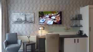 A modern hotel room with a dining area, a TV mounted on the wall, a chair, a small table, and a cabinet. - Strand Hotel Dangast (Varel)