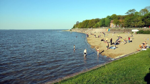 A beach with people swimming and sunbathing, a grassy area, and a building in the background. - Pension an Bord (Varel)