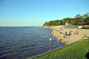Ein Strand mit badenden und sonnenden Menschen, einer Grünfläche und einem Gebäude im Hintergrund.