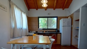 A small kitchen with a wooden table and chairs, a refrigerator, and a window with a view of greenery. - Cabanas Nagual (El Hoyo)