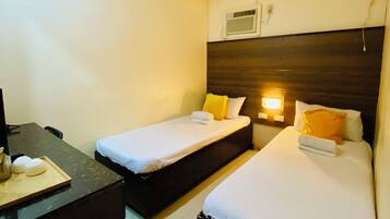 A hotel room with two single beds, a desk with a chair, a trash bin, and a wall-mounted air conditioning unit.