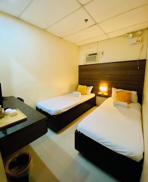 A hotel room with two single beds, a desk with a chair, a trash bin, and a wall-mounted air conditioning unit.
