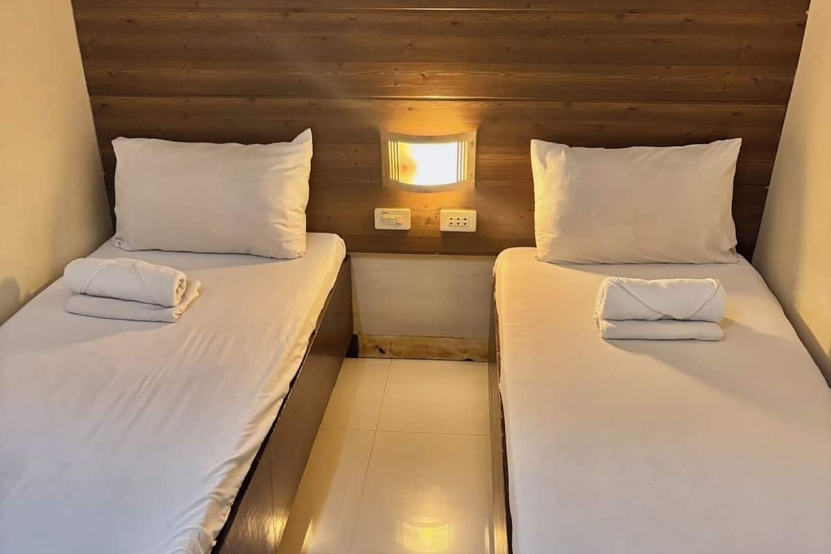 A room with two beds, each with white linens and pillows, a small wall-mounted light, and a wooden headboard.
