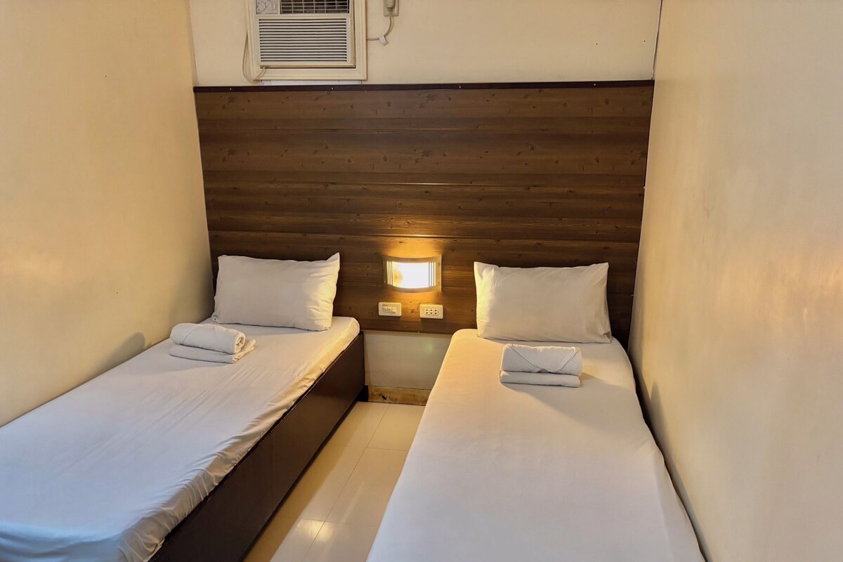 A small, cozy room with two beds, wooden headboard, bedside lamps, and a window with blinds.