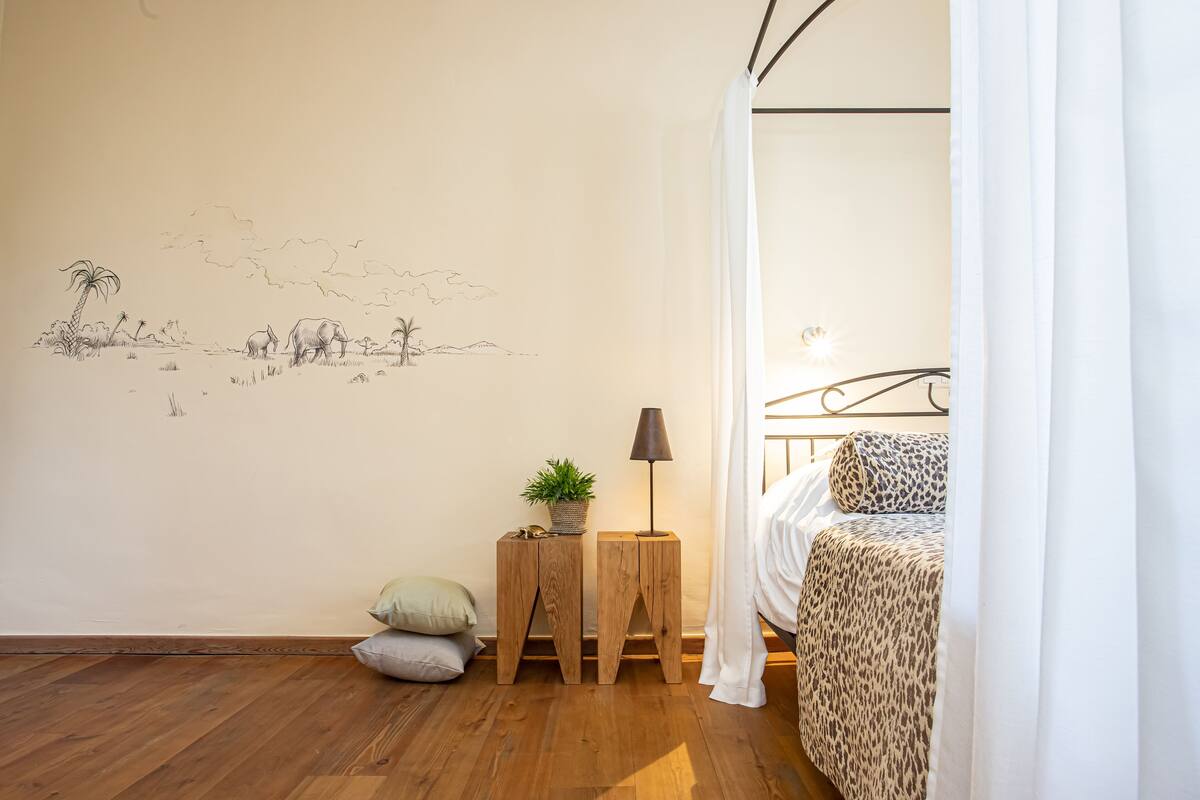 A bedroom with a bed, a nightstand with a lamp, a small plant, and a wall mural.