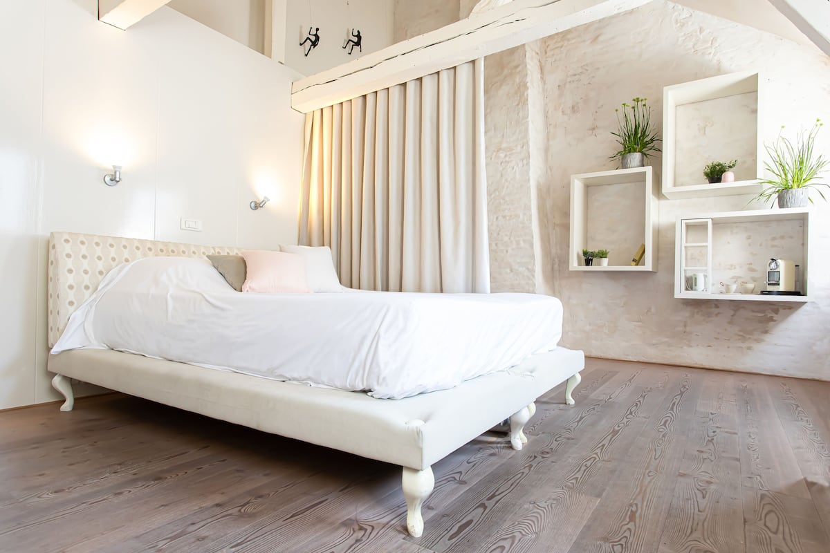 A spacious bedroom with a large bed, wooden flooring, a sloped ceiling, and built-in shelving with plants.
