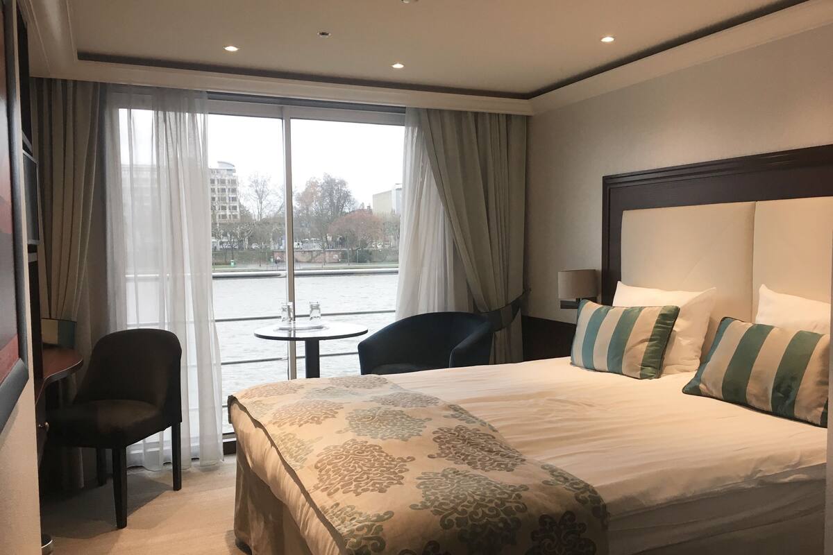 Une chambre d’hôtel avec un lit, une chaise, une petite table et une vue sur une étendue d’eau, visible à travers de grandes fenêtres.
