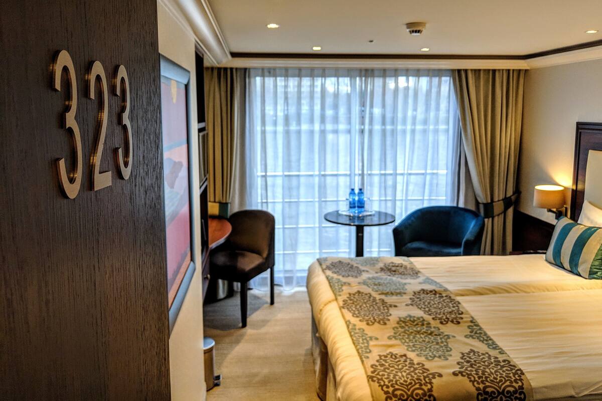 Une chambre d’hôtel avec un lit, une chaise, une petite table avec des bouteilles d’eau et une grande fenêtre avec des rideaux.