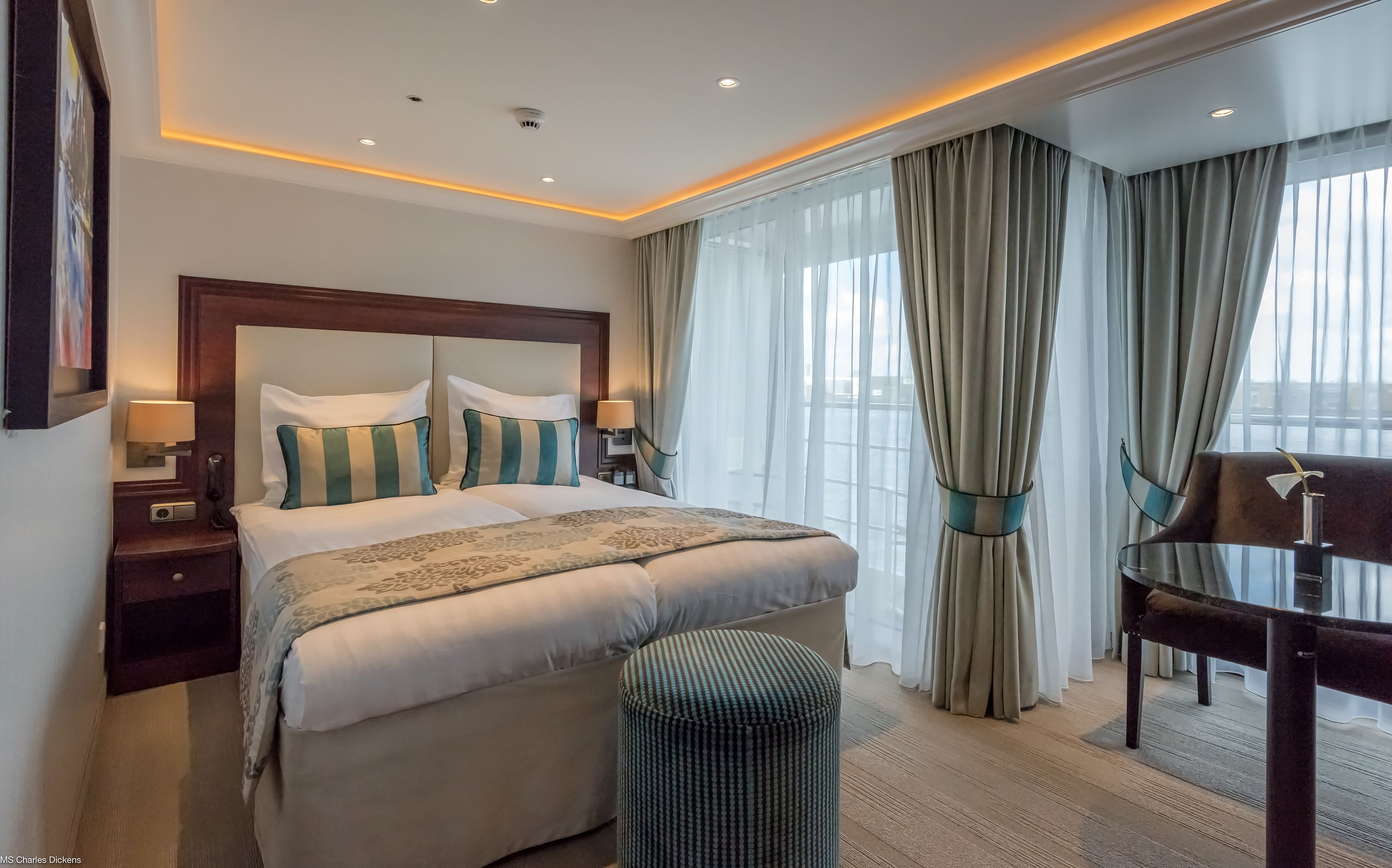 Ein Hotelzimmer mit einem großen Bett, zwei Nachttischen inklusive Lampen, einem kleinen runden Hocker, einem Schreibtisch mit Stuhl und einem großen Fenster mit Vorhängen.