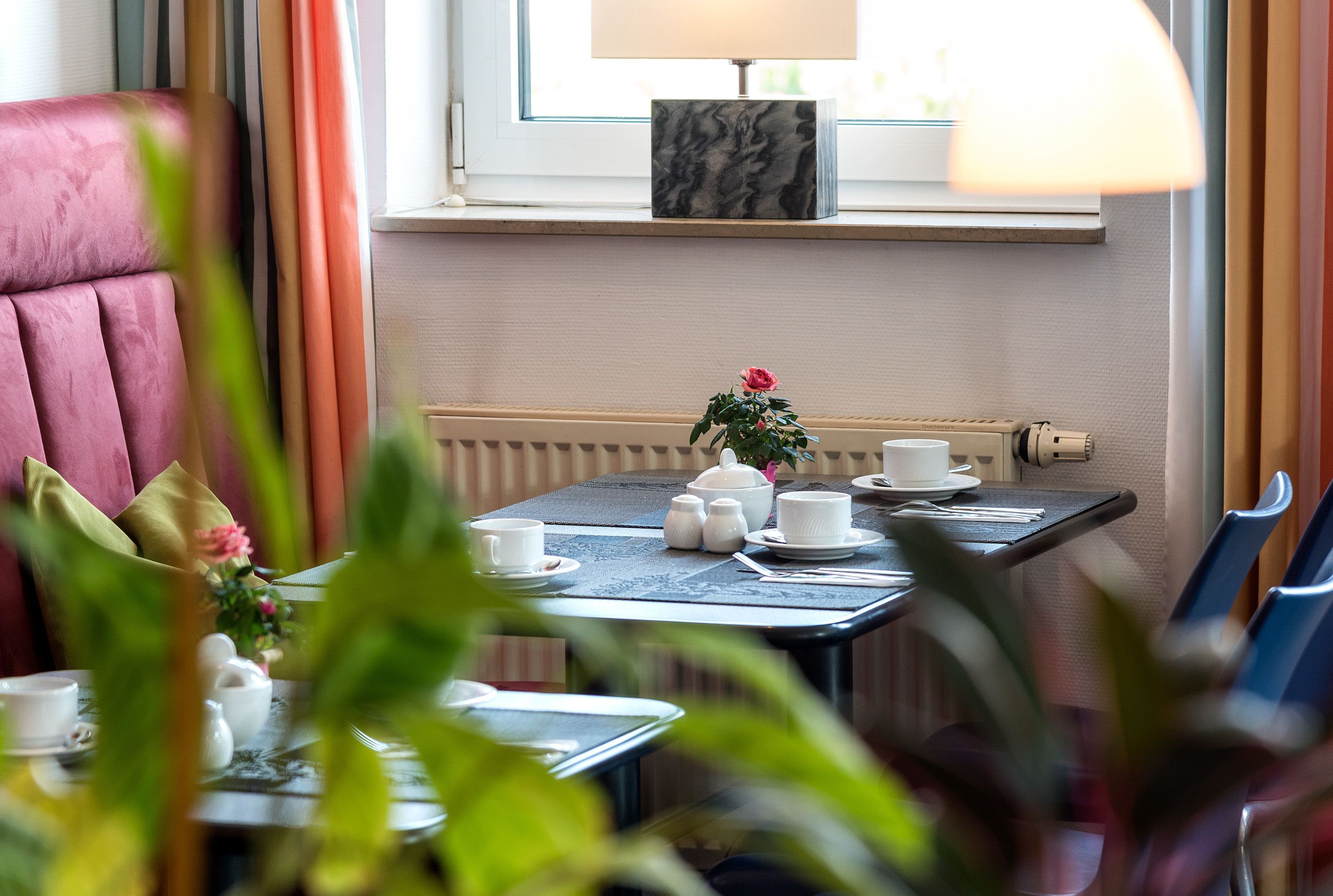 Comedor con una mesa dispuesta para dos, con tazas de té blancas, platillos, una tetera y un azucarero. Se ve una ventana con vistas al exterior, un radiador y una lámpara de mesa en el alféizar.