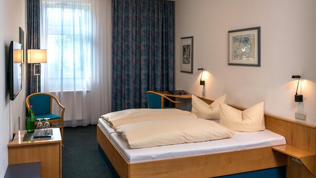 Une chambre d’hôtel comprenant un lit, un bureau, une chaise, une lampe de bureau, une table de chevet et une fenêtre avec des rideaux.