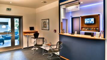 A reception area with a reception desk, two chairs, a small table with a magazine, a framed picture on the wall, and a television screen above the desk.