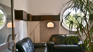 A modern living room with two black leather sofas, a large floor lamp, and a round window offering a view of greenery. - Median Hotel Garni (Wernigerode)