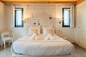 Un dormitorio con una cama grande, dos mesitas de noche, una silla y dos ventanas con cortinas.