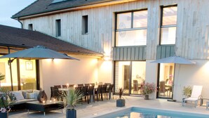 A swimming pool with a wooden deck, outdoor seating, and a building with large windows and a sloped roof. - Ome Sweet Home (Laubressel)